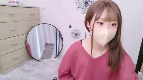 Media: Video of an Asian woman with long brown hair, wearing a pink sweater and a face mask, sitting on a fluffy white rug in a minimalist room with a dresser, mirror, and black dandelion wall art.