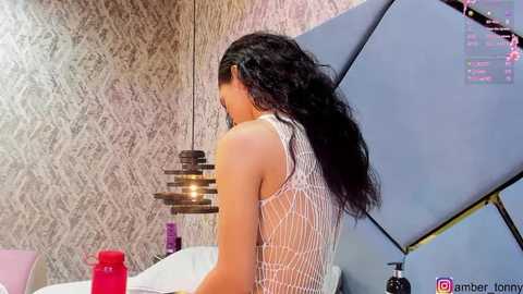 Media: Video of a woman with long, black hair in a white, see-through lace top, standing in a modern room with a gray, textured wallpaper, and a blue, abstract art piece on the wall.