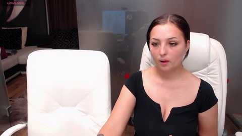 Media: Video of a fair-skinned woman with brown hair, wearing a black top, seated in a white leather chair, in a dimly lit office with a computer monitor and a window.