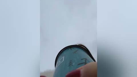 Media: A close-up video of a person's hand holding a blue glass bottle with a white label featuring black text, partially obscured by the bottle's curvature. The background is out of focus, creating a blurred, white, and slightly grainy effect.