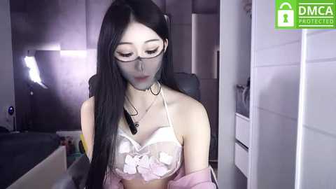 Media: Video of an Asian woman with long black hair and glasses, wearing a see-through lace bra, sitting in a dimly-lit room with white walls and a bathroom cabinet.