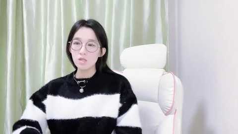 Media: Video of a young Asian woman with pale skin, straight black hair, and round glasses, wearing a black-and-white striped sweater, seated on a white leather chair against green curtains.