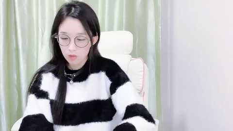 Media: Video of an East Asian woman with long black hair, wearing large round glasses and a black-and-white striped sweater, sitting on a white chair against a green curtain background.