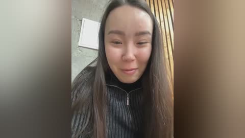 Media: Video of an Asian woman with long black hair, wearing a black pinstriped shirt, looking introspective in a dimly lit room with a concrete wall and wooden paneling.