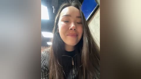 Media: Video of a young woman with long brown hair, wearing a black and gray striped sweater, standing in a dimly lit, modern indoor setting with white walls and a blue sky visible through a window.