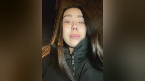 Media: A close-up video of a young woman with long, straight brown hair, wearing a black puffer jacket, smiling and blowing a kiss. The background is blurred and dark.