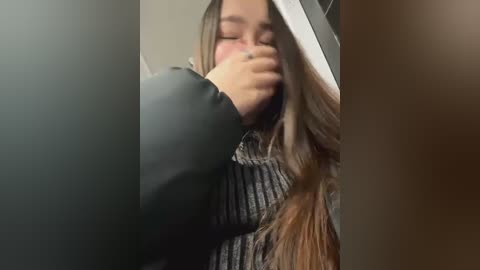 Media: A video captures a young woman with long brown hair, wearing a dark sweater, covering her face with her hand, possibly in a moment of embarrassment or discomfort. The background is blurred and nondescript.