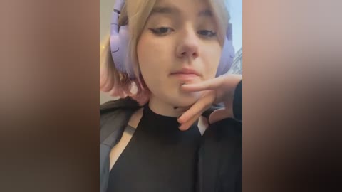 Media: Video of a young woman with pale skin, light blonde hair, wearing large purple headphones and a black shirt, resting her chin on her hand in a casual, introspective pose.