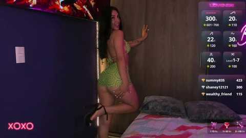 Media: Video of a curvaceous woman with long dark hair, wearing a lime green, backless lingerie set, standing provocatively near a bed, in a dimly lit bedroom.