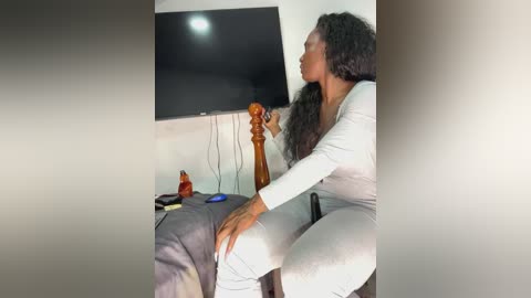 Media: A video shows a Black woman with long, curly hair in a gray jumpsuit, sitting on a gray couch, holding a remote, in a dimly lit room with a large, flat-screen TV on the wall.