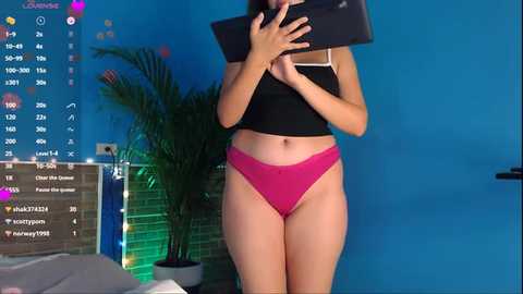 Media: Video of a light-skinned woman with a slim build, wearing a black crop top and pink panties, holding a smartphone, in a blue-walled room with a green screen, plants, and a calendar.