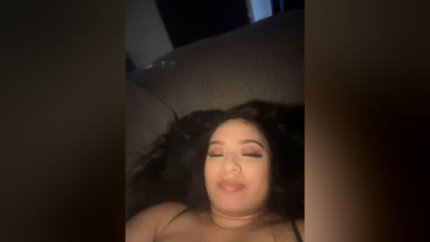 Media: Video of a woman with medium skin tone and curly dark hair lying on a dark leather couch, eyes closed, lips pursed, wearing a beige top.