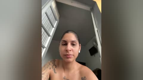 Media: A video of a topless Latina woman with medium skin tone, medium build, and short dark hair, standing indoors. She has tattoos on her shoulders and is wearing small hoop earrings. The background shows a dimly lit room with closed white blinds and a mounted TV.