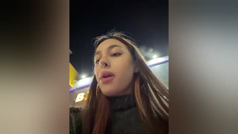 Media: A video of a young Asian woman with long brown hair and a neutral expression, taken at night in an urban setting with bright lights and a dark sky background.