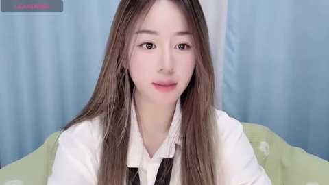 Media: Video of a young East Asian woman with long, straight, brown hair, light skin, and subtle makeup, wearing a white blouse and black tie, sitting against a light blue curtain background.