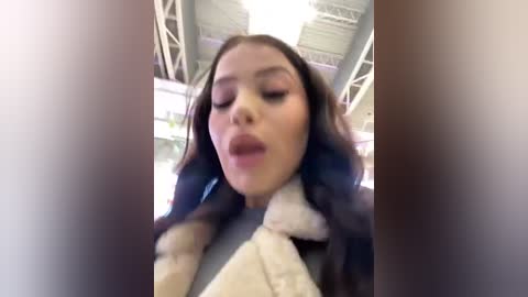 Media: A blurry video of a young woman with long, wavy hair and closed eyes, wearing a fur-trimmed coat, standing indoors near metal beams.