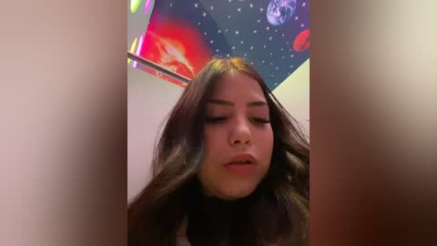 Media: A video of a young Latina woman with long, dark hair, wearing a black shirt, standing in a room with a colorful, space-themed mural on the wall.