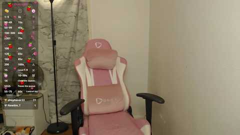 Media: Video of a pink gaming chair with white accents and armrests, positioned in a corner against a beige wall. On the left, a black lamp with a white shade stands beside a gray, patterned curtain.