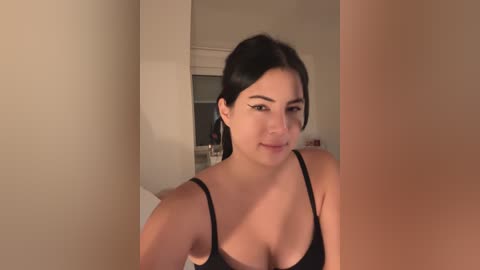 Media: Video of a young Latina woman with medium skin tone, dark hair, and black bra, standing in a beige-walled bathroom, looking into a mirror.