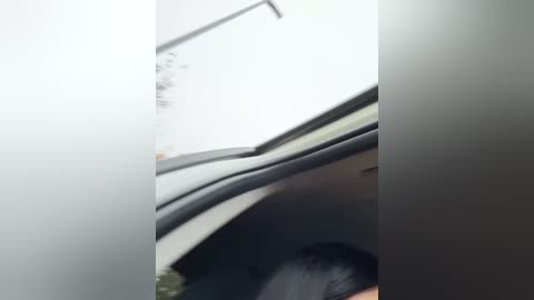 Media: Video of a person with long black hair sitting in a car, head slightly turned, with a blurred background of a white wall and a partially open window.