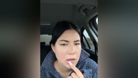 Media: Video of a young woman with dark hair, wearing a blue sweater, sitting in a car. She has a finger to her lips, indicating silence. The background shows car seats and windows.