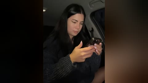 Media: Video of a young woman with long black hair, fair skin, and closed eyes, wearing a dark knit sweater, holding a smartphone, sitting in a car.