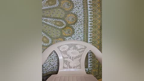 Media: A video featuring a cream-colored plastic chair with intricate floral patterns, placed against a backdrop of a vibrant, ornate tapestry with shades of green, yellow, and white. The setting suggests a traditional or cultural interior.