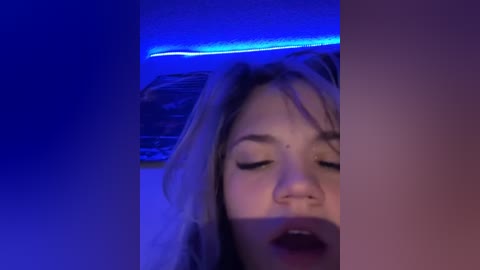 Media: Video of a close-up of a woman's face, with closed eyes and parted lips, illuminated by blue light. Her skin is light, and she has blonde hair.