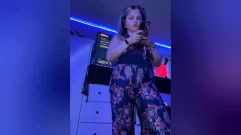 Media: A video of a young woman with light skin and long brown hair, wearing a dark floral jumpsuit, taking a selfie in a dimly lit room with blue LED strip lighting.