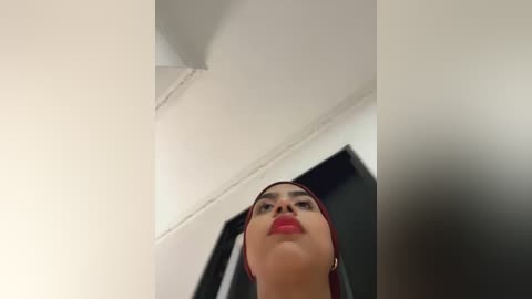 Media: A low-angle video of a young woman with fair skin and dark hair, wearing a red hijab and bold red lipstick, standing in a minimalist white room with a black TV in the background.