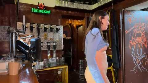 Media: Video of a pregnant woman in a dimly lit bar, wearing a blue shirt and yellow pants, standing next to a Heineken sign.