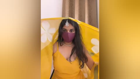 Media: Video of a woman with long dark hair, wearing a bright yellow saree, purple mask, and floral headpiece, standing in front of a yellow screen with white flowers.