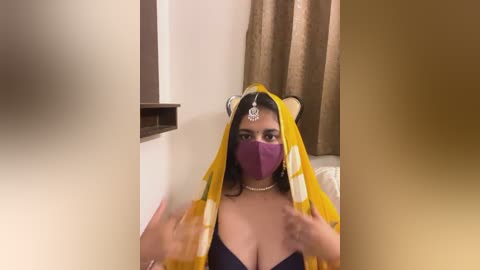 Media: Video of a young woman with medium skin tone, wearing a yellow and white sari, purple mask, and silver headpiece, standing in a bedroom with beige curtains.