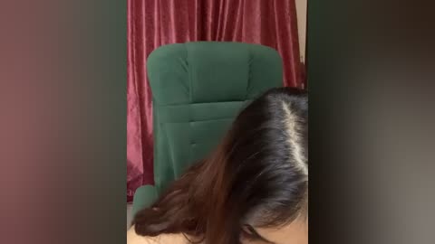 Media: A video showing a person with dark hair sitting in a green velvet armchair against a red velvet backdrop. The person\u2019s head is tilted forward, revealing their shoulder and back.