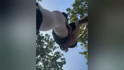 Media: Video of a woman with fair skin and medium build, wearing a black tank top and white leggings, bending over in a forest with green trees and a clear blue sky.