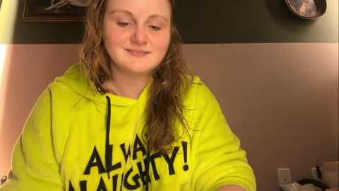 Media: Video of a young woman with wet, shoulder-length brown hair, wearing a bright yellow hoodie with \"ALWAYS!\" and \"NAUGHTY!\" in bold black letters. She smiles softly, standing indoors against a beige wall with a framed picture and clock above.