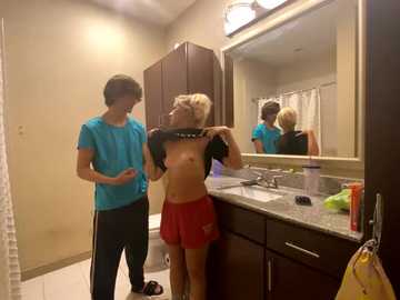 Media: Video of a blonde woman in a bathroom, lifting her shirt, exposing her breasts, while a man in blue shorts watches, both in a modern bathroom with a granite countertop and mirror.