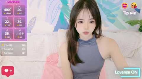 Media: Video of a young Asian woman with long black hair, wearing a tight, sleeveless blue dress, sitting on a bed with pink sheets. Background features a colorful abstract painting.