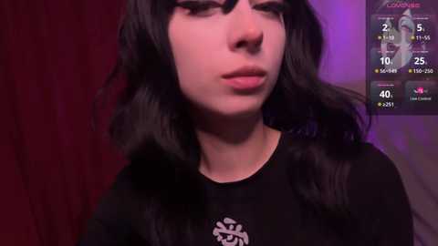 Media: A close-up video of a young woman with pale skin, long black wavy hair, and dark makeup, wearing a black shirt with a white emblem, against a dim, red background.