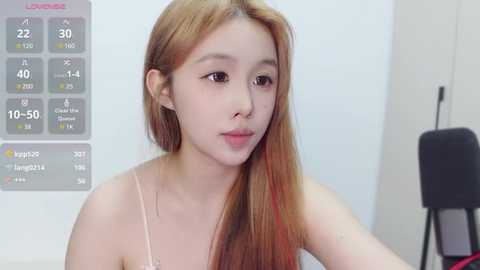 Media: Video of an Asian woman with long, straight, reddish-brown hair, wearing a light-colored spaghetti-strap top, standing in a minimalist room with a weather forecast displayed on the left.