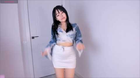 Media: Video of an Asian woman with straight black hair and fair skin, wearing a white denim jacket and high-waisted white shorts, standing in a white-walled room with a white door.