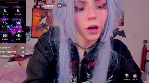 Media: Video of a young woman with pale skin, long blue hair, and dramatic makeup, wearing a black jacket with metal studs, in a bedroom with a colorful cat poster and gaming interface displayed.