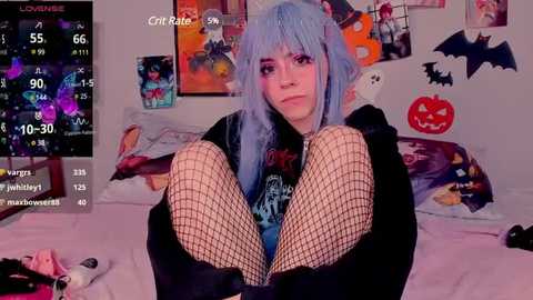 Media: Video of a young woman with blue hair, dressed in black fishnet tights, sitting on a bed in a room adorned with Halloween decorations, including a pumpkin and bat stickers.