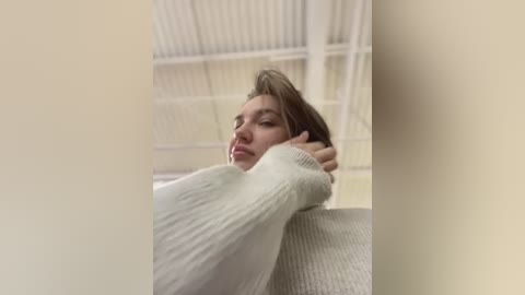 Media: A video of a young woman with light skin, brown hair, and a relaxed expression, wearing a light grey, ribbed sweater, leaning against a white metal structure, possibly a warehouse, with blurred, industrial background.