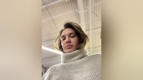 Media: Video of a young woman with fair skin, wearing a cream-colored ribbed turtleneck sweater, standing in a modern, industrial-style room with white metal beams and fluorescent lighting. Her short, light brown hair is slightly tousled.