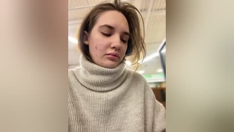 Media: Video of a young woman with fair skin and straight brown hair, wearing a cream turtleneck sweater, with a small pimple on her cheek. Background features a beige ceiling and blurred, modern interior.