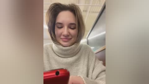 Media: A video of a young woman with light skin and short, layered brown hair, wearing a cream-colored turtleneck sweater, smiling while looking at a red smartphone. The background shows a blurred, indoor setting with a metallic surface and ceiling lights.