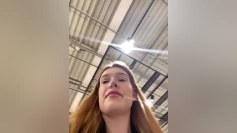 Media: A video of a young woman with long, wavy, light brown hair, looking up from a low angle, captured indoors under a high, industrial ceiling with exposed beams and fluorescent lights.