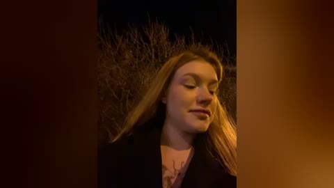 Media: Video of a young woman with long blonde hair, wearing a black coat, standing in a dark, barren field at night, illuminated by warm light.