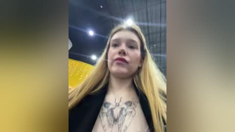 Media: A video of a young, fair-skinned woman with long blonde hair, wearing a black jacket, with a large tattoo of three bats on her chest, standing in a dimly lit, modern room with yellow and black accents.